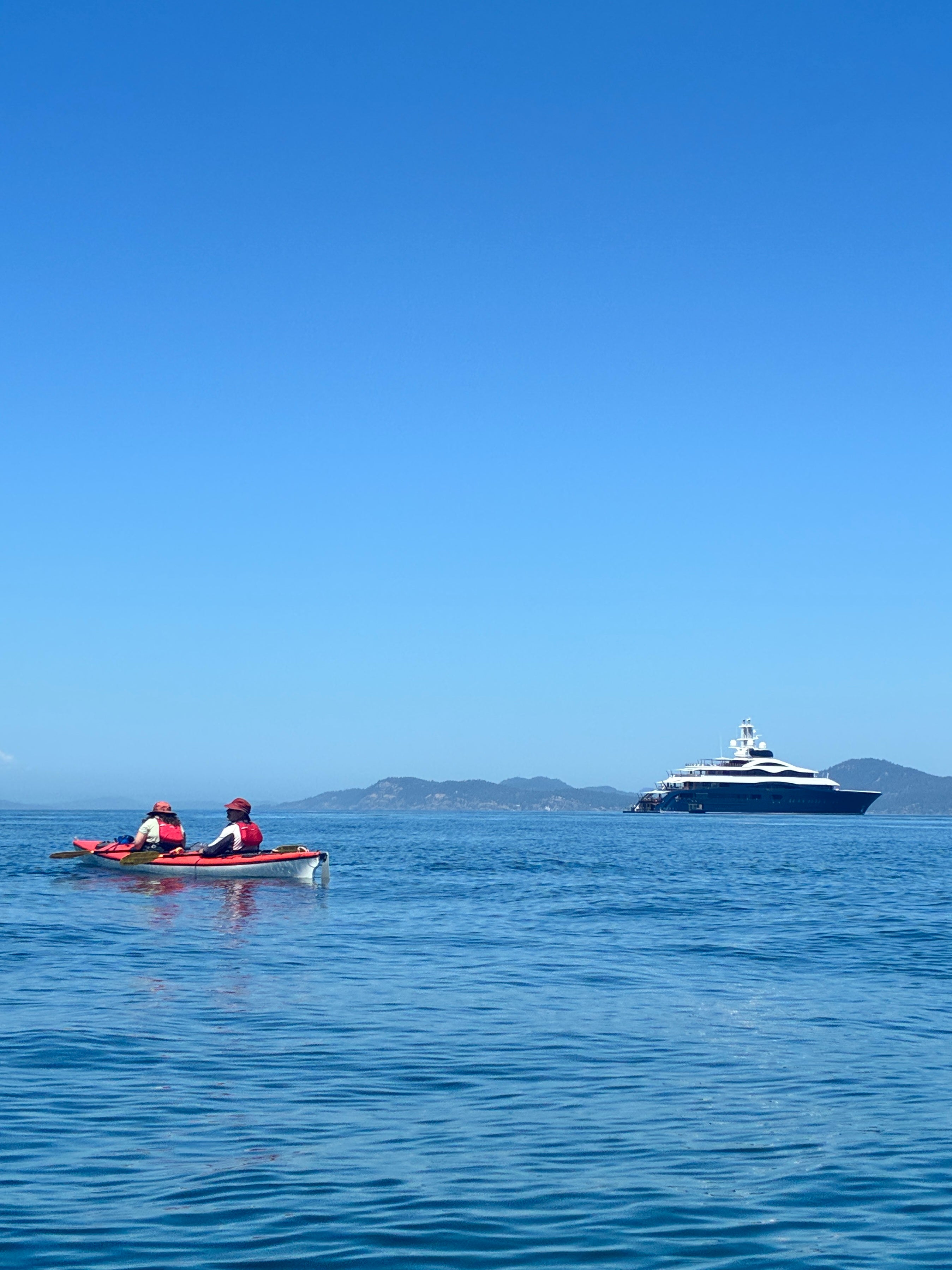 Two people kayaking in a red kayak near a distant white yacht on calm blue sea under clear sky