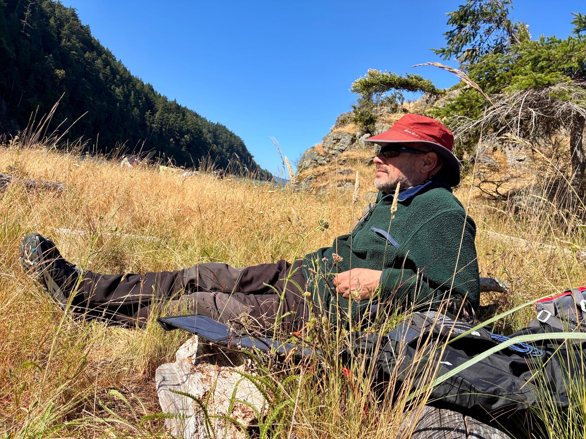 Nicholas Pinter reclining in grassy island meadow wearing red hat and green sweater beside backpack