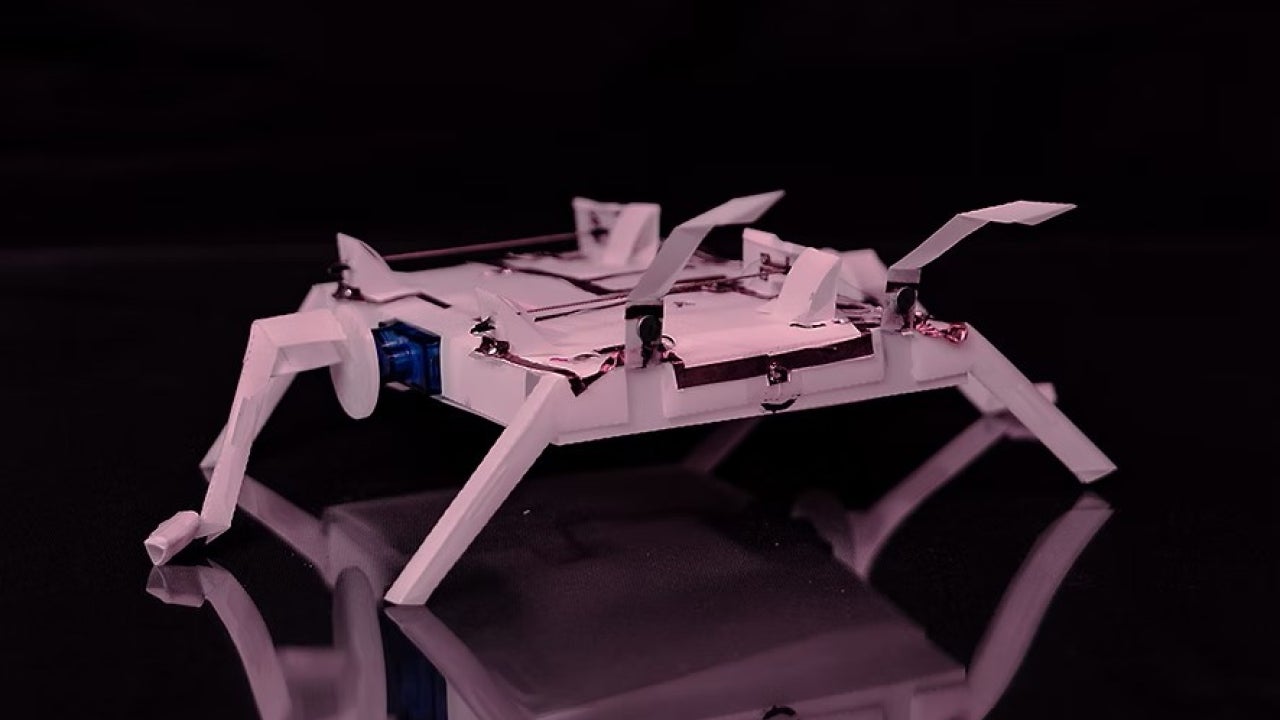 A robotic device resembling a spider, with four legs and a flat body, set against a dark background.