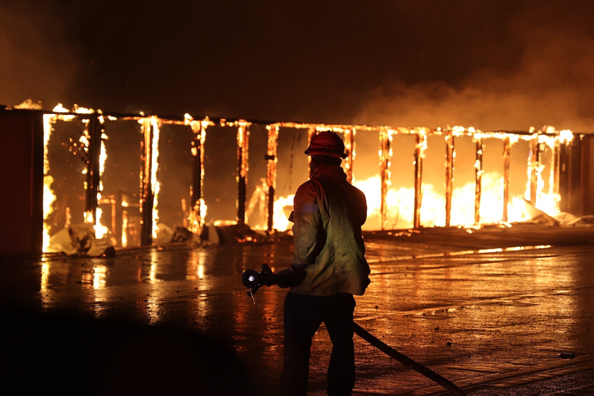 Silhouetted firefighter holding a hose before a row of blazing buildings. (Courtesy CAL FIRE)