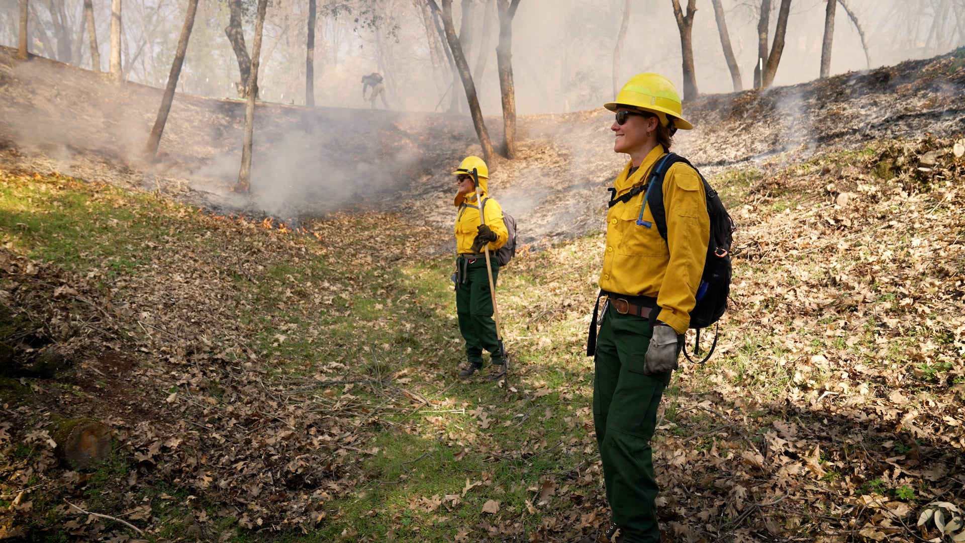 Two women in yellow firefighting shirts and helmets stand in meadow with light smoke in background during prescribed burn.