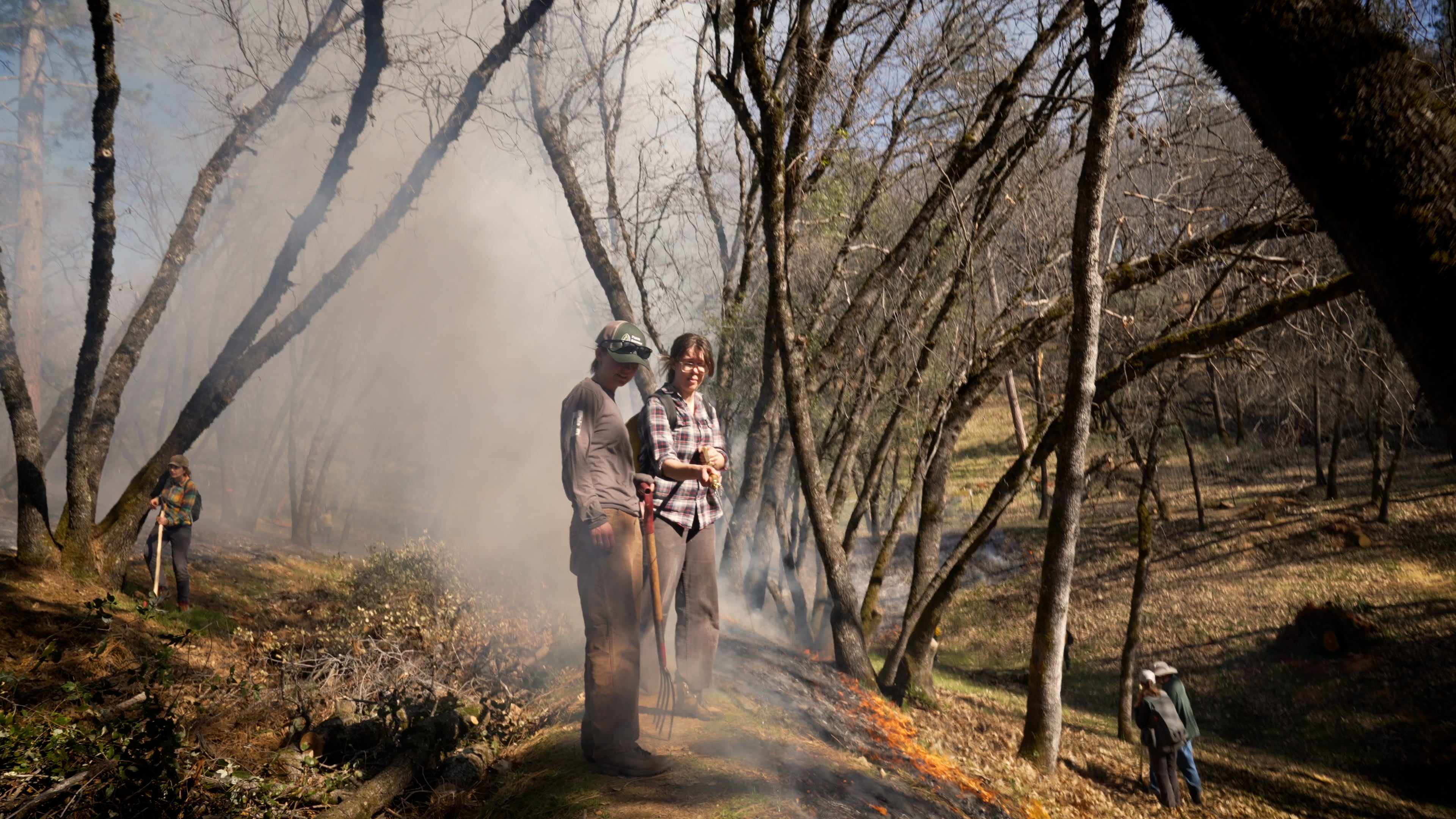 Two women on a hillside with smoke behind them during prescribe ffire