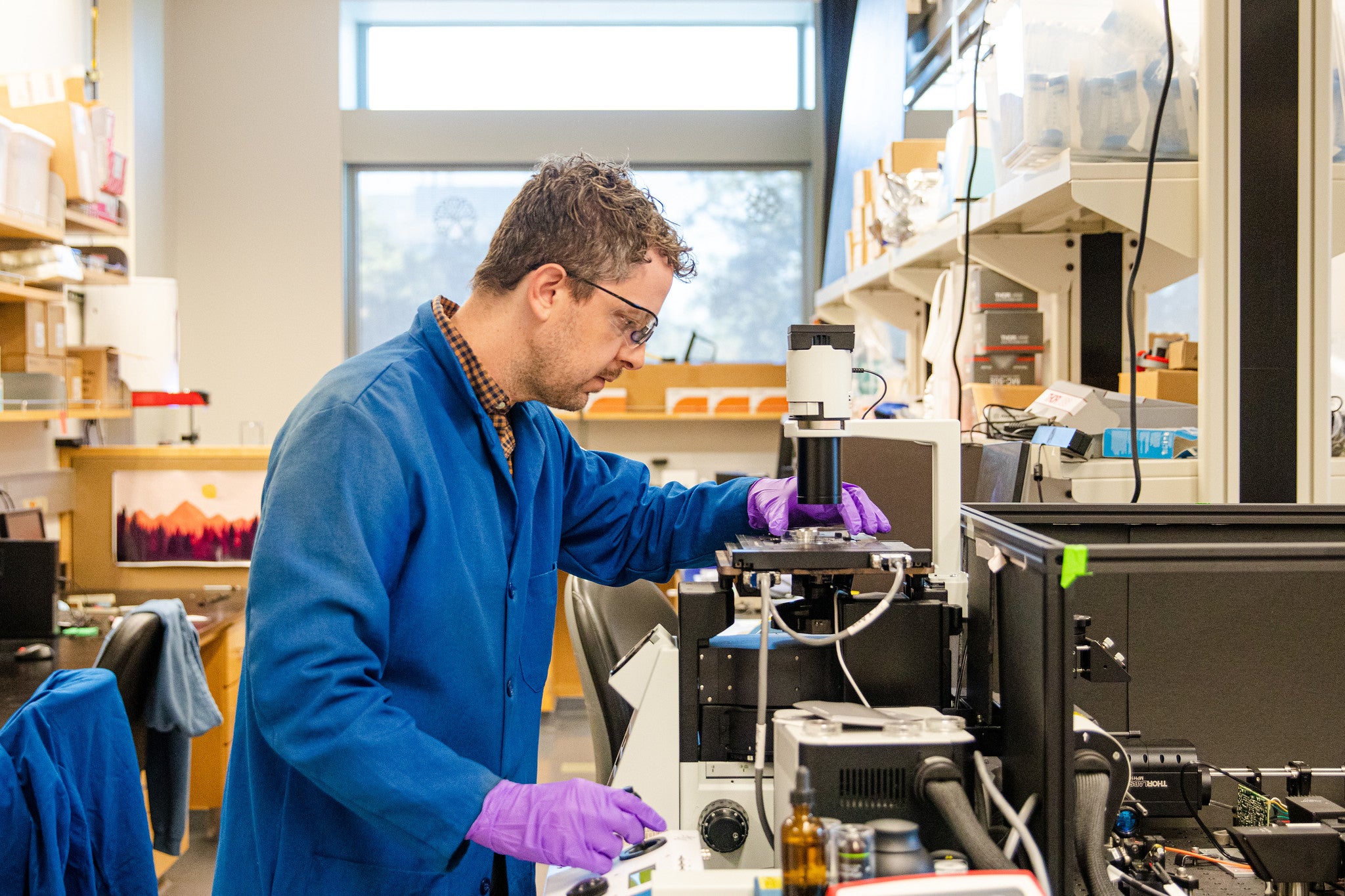 Randy Carney in blue lab coat and purple gloves using a microscope in a lab