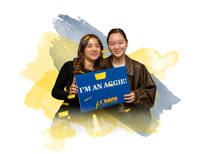 Two young women smile while holding a sign that says "I'M AN AGGIE!" against a colorful background.