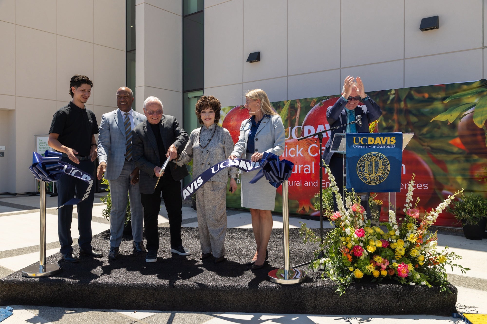 Seven people at an outdoor ribbon-cutting, holding giant scissors, banner and floral display in front of a new building. From left: UC Davis student Jose Gomez, Chancellor Gary S. May, philanthropists Stewart and Lynda Resnick, Dean Ashley M. Stokes, and Vice Chancellor Shaun Keister celebrate the opening of the Resnick Center for Agricultural Innovation on Tuesday, May 19, 2026. (Courtesy of The Wonderful Company)