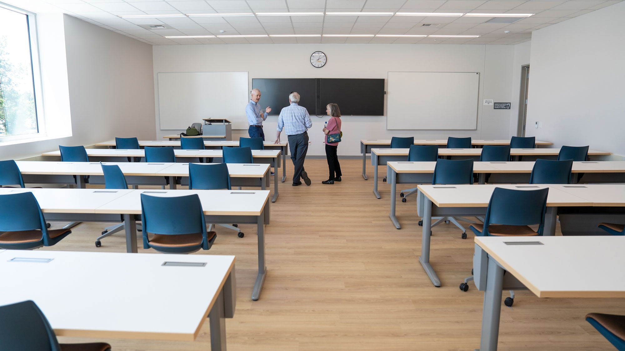 Classroom with rows of white desks and blue chairs, three people talking at front
