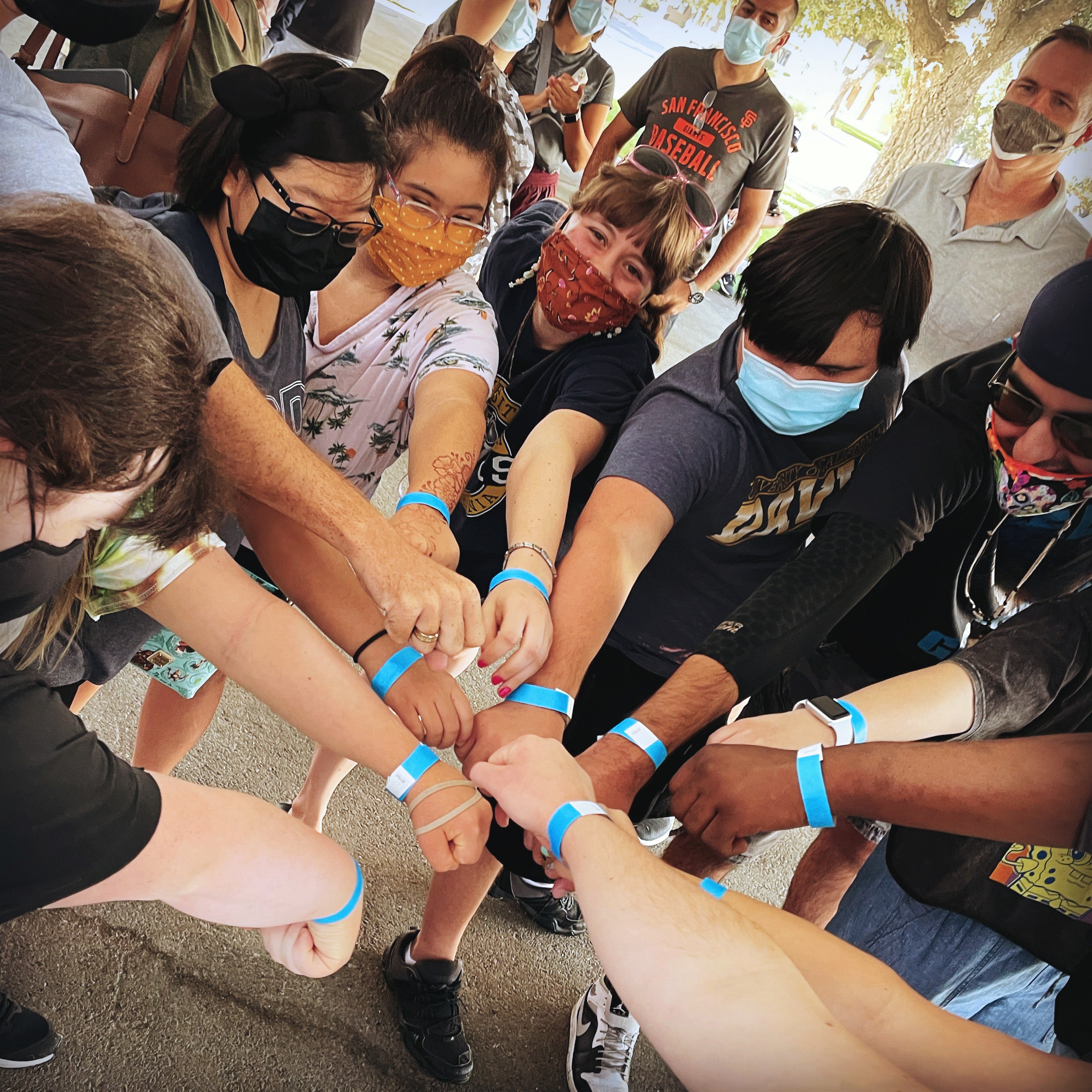 A group of people reach their hands into the center of a the circle and show off blue wristbands