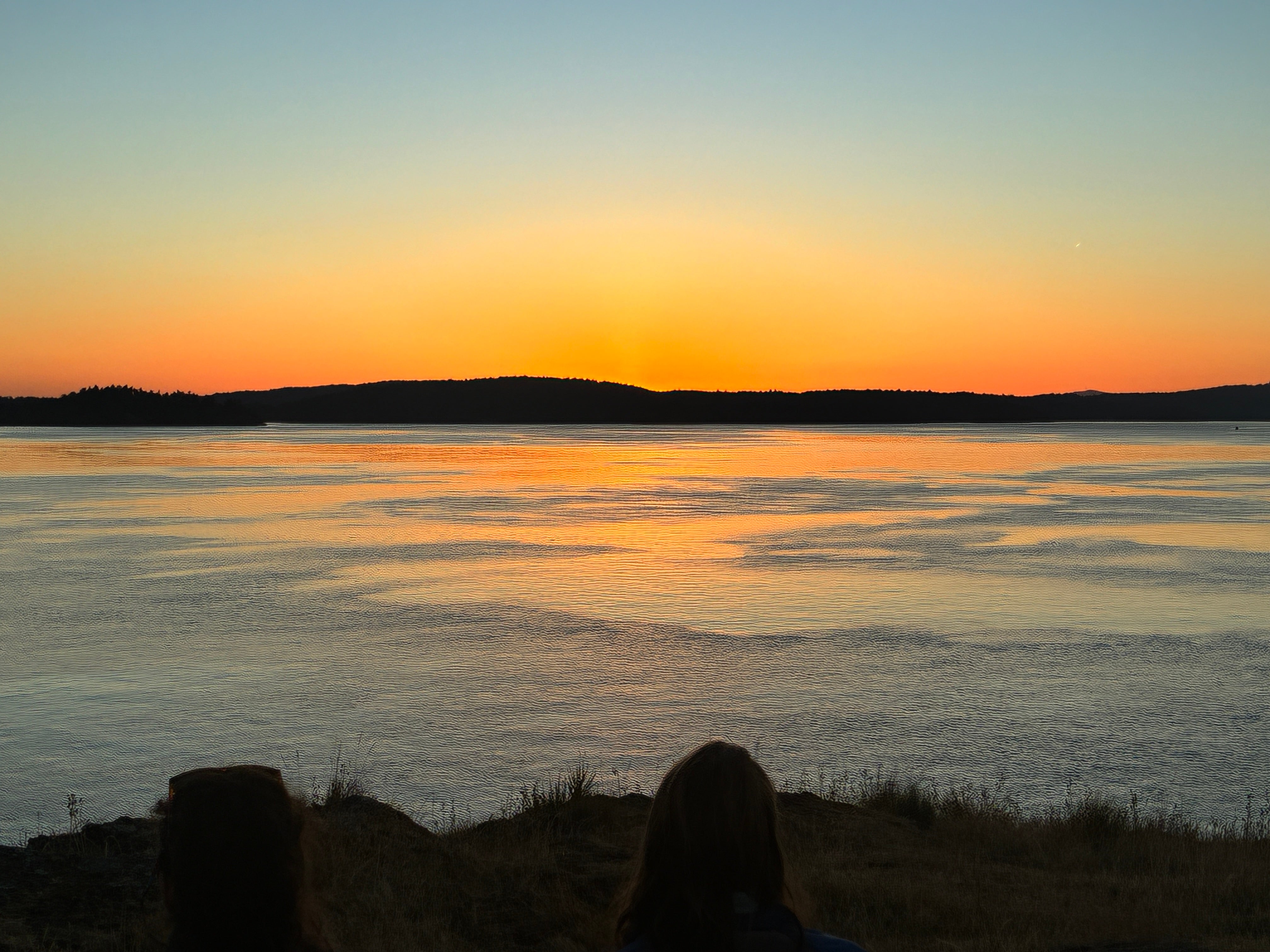 Orange sunset over calm water with two silhouetted people on a grassy bluff