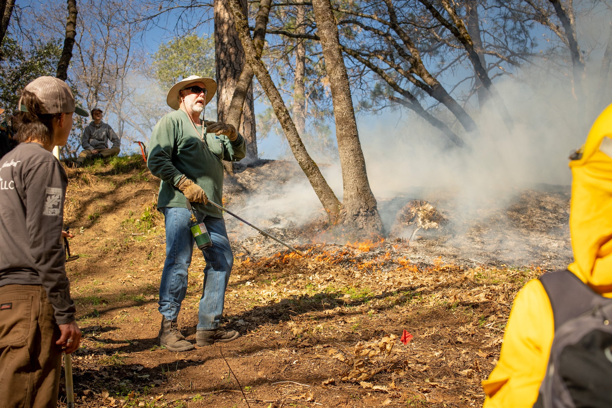 Man with beard and hat talks to group in wooded clearing during a prescribed burn