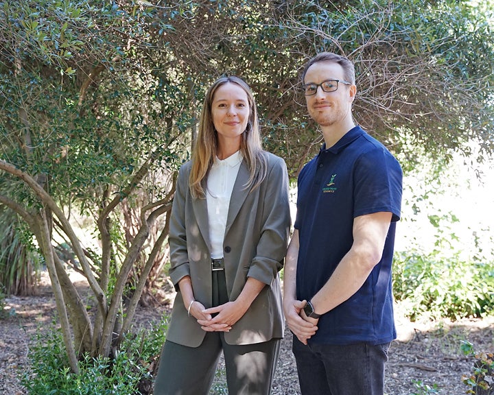 Young woman in gray blazer and man in navy polo standing in a shaded garden, photo