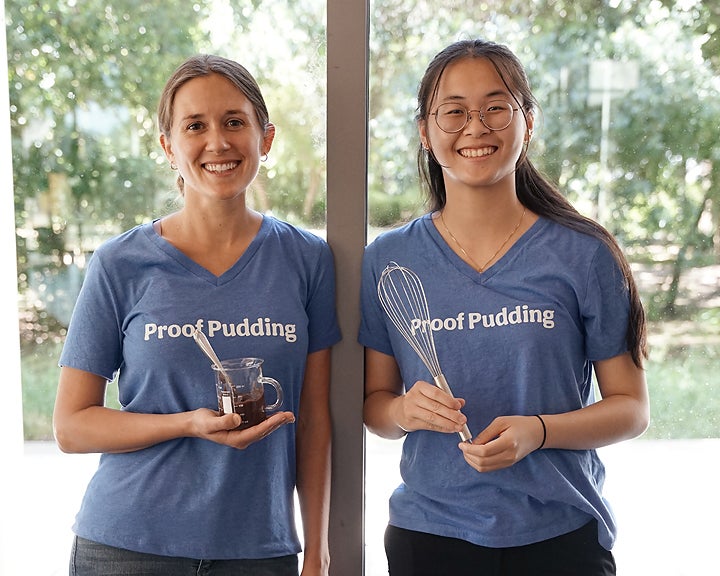 Two smiling women in matching 'Proof Pudding' tees holding a glass jar and whisk outdoors