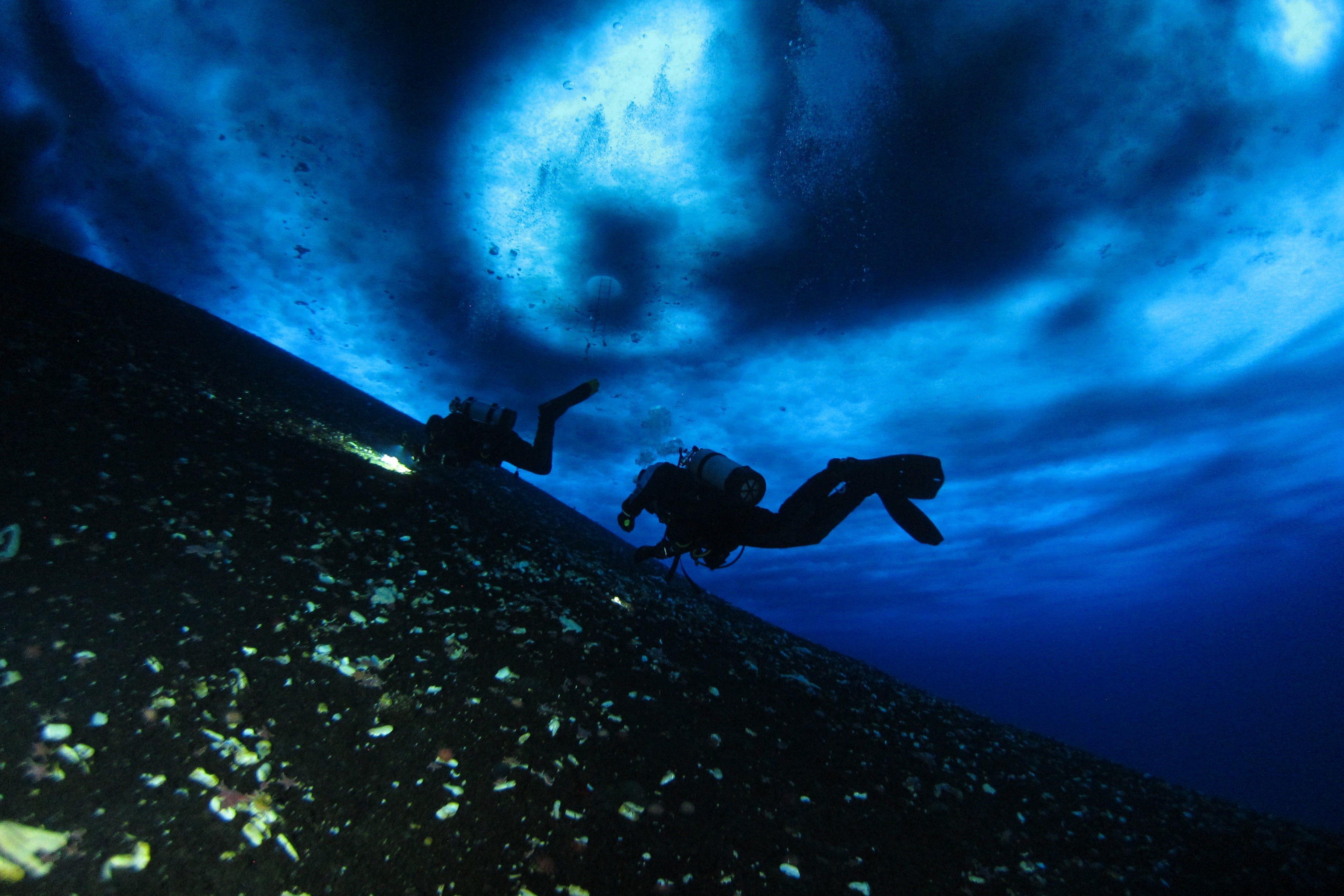 Divers swim under ice in Antarctica in otherworldy landscape of swirling blues above