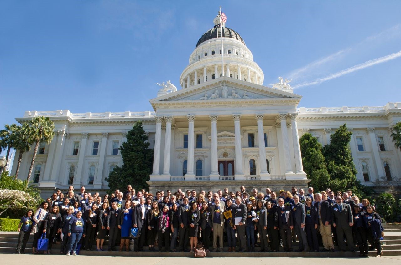 A large group of students and staff members in business attire pose on the steps of the California State Capitol.