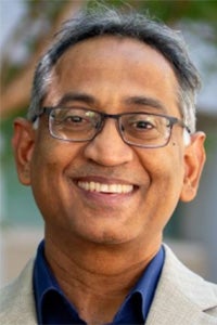 Smiling man wearing glasses, tan blazer and navy shirt, outdoor portrait