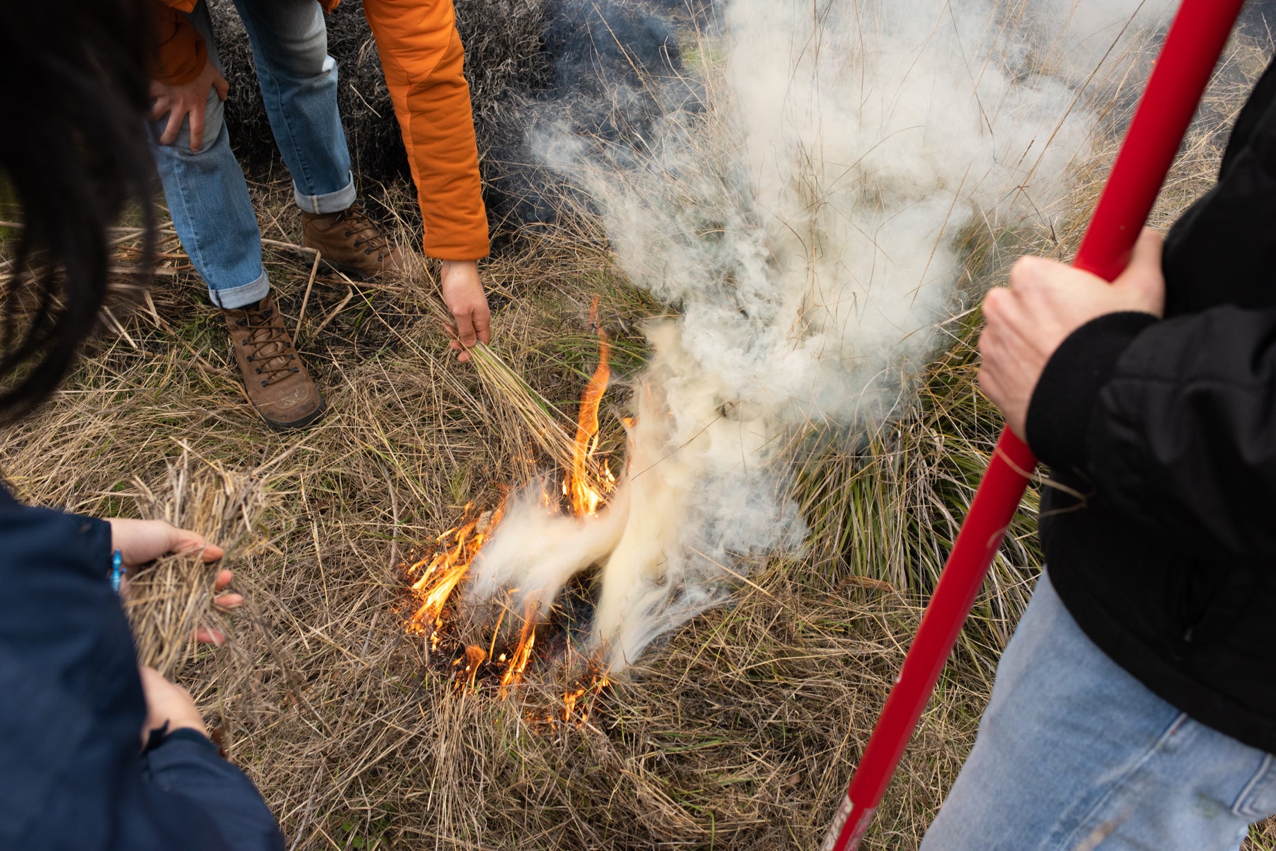 a small flame ignites dried grass as three people with red-handled tools are visible from waist down.