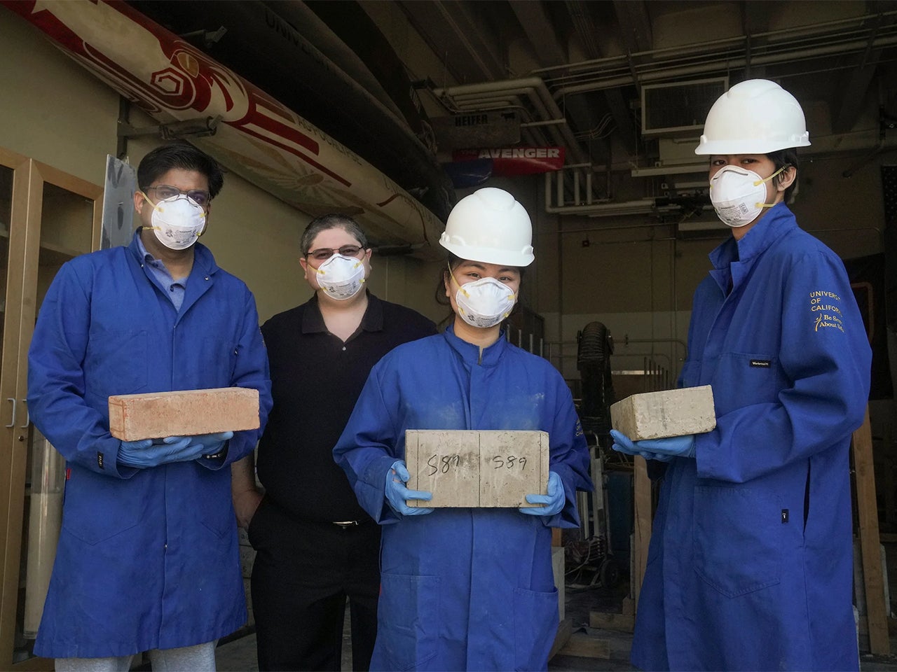 Four people in blue construction suits and white hard hats and face masks pose with earth blocks