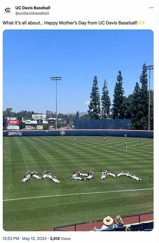 Full tweet text: "What it's all about... Happy Mother's Day from UC Davis Baseball! 🌹" Image description: The image shows the baseball field at UC Davis. The players are lying down on the outfield grass arranged to spell out "MOM" as a Mother's Day greeting. There are tall trees and stadium lights in the background, along with advertisements on the outfield walls. In the foreground, there appears to be a person wearing a hat sitting in the stands or viewing area.
