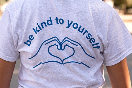 Grey shirt with blue text: Be kind to yourself, illustration of hands folded in heart shape.
