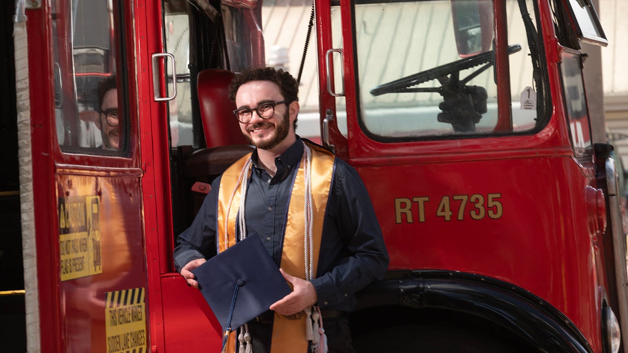 Smiling young man in graduation gown and gold stole holding cap and standing in front of a red vintage bus, smiling beside red vintage bus.