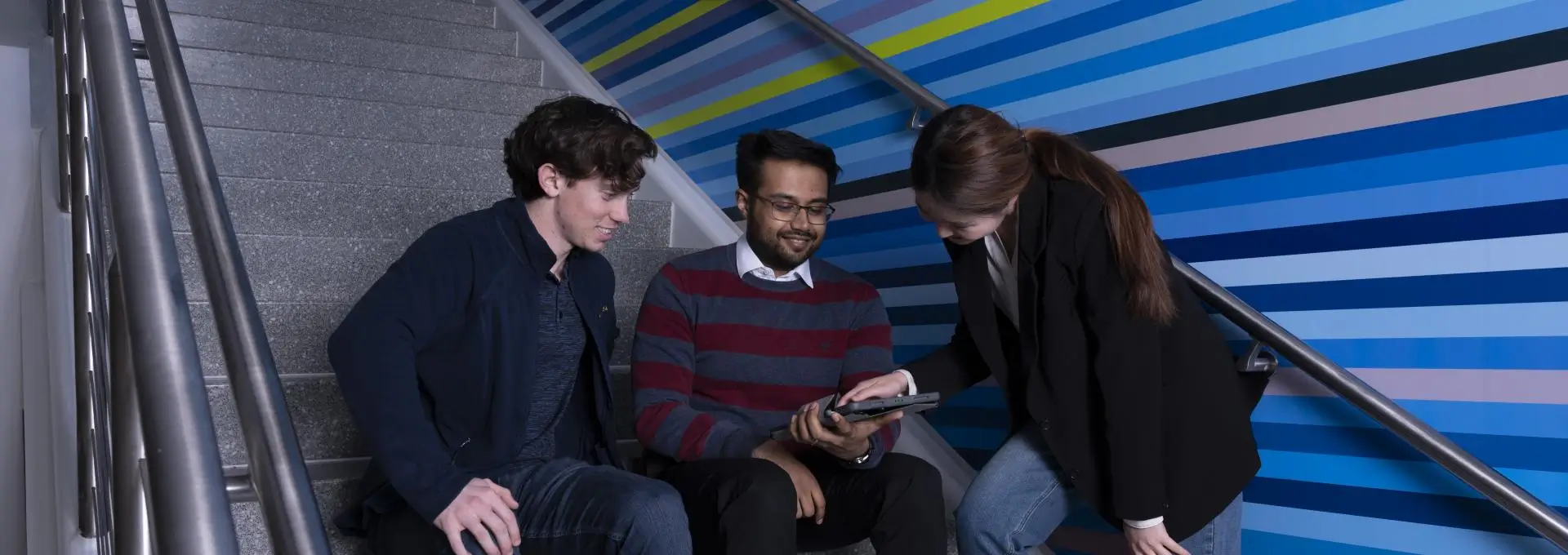 three students sitting on steps talking and looking at a device