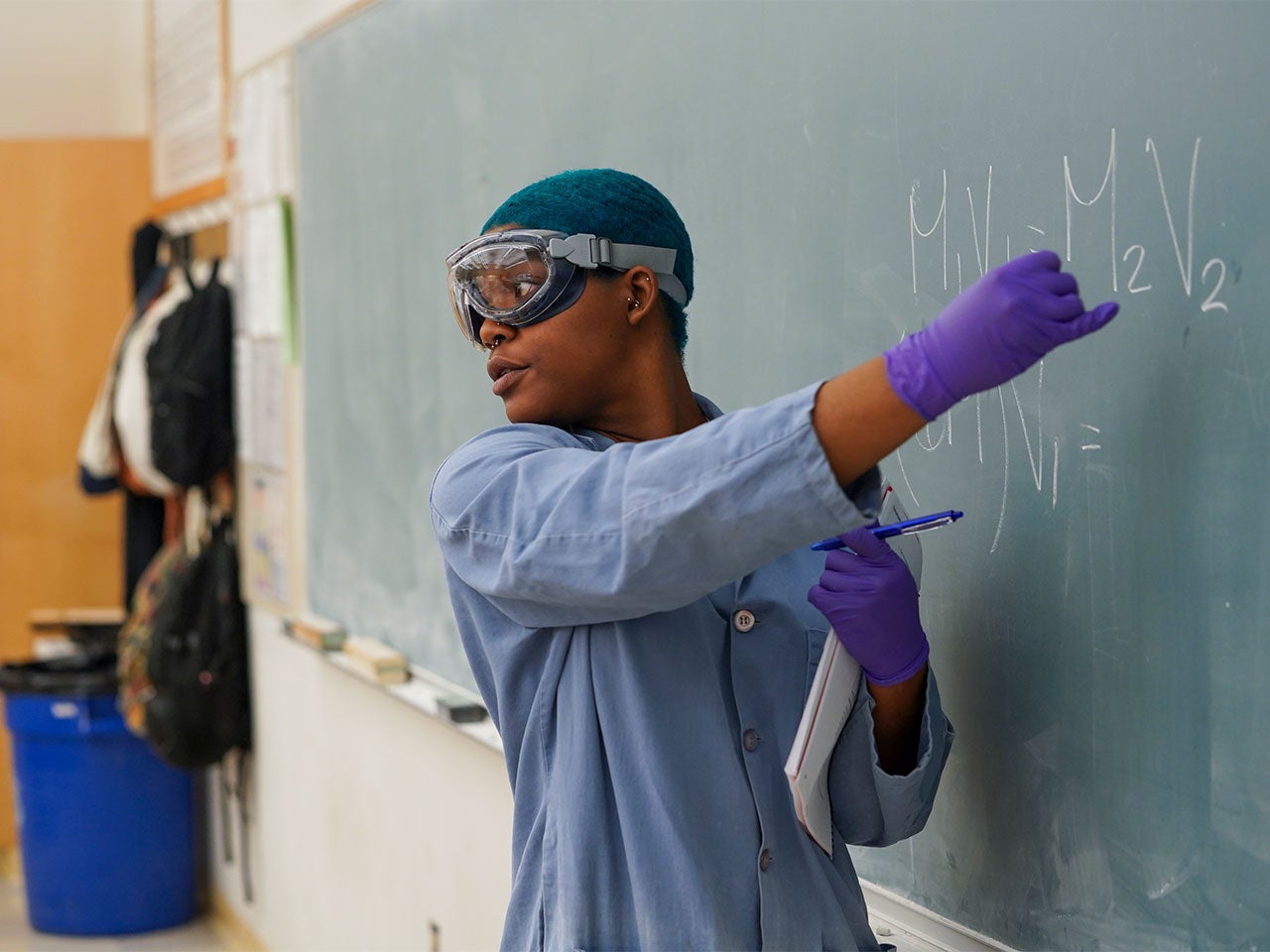 A UC Davis teaching assistant with short blue hair and wearing a blue lab coat points to a chemical equation on a blackboard. 