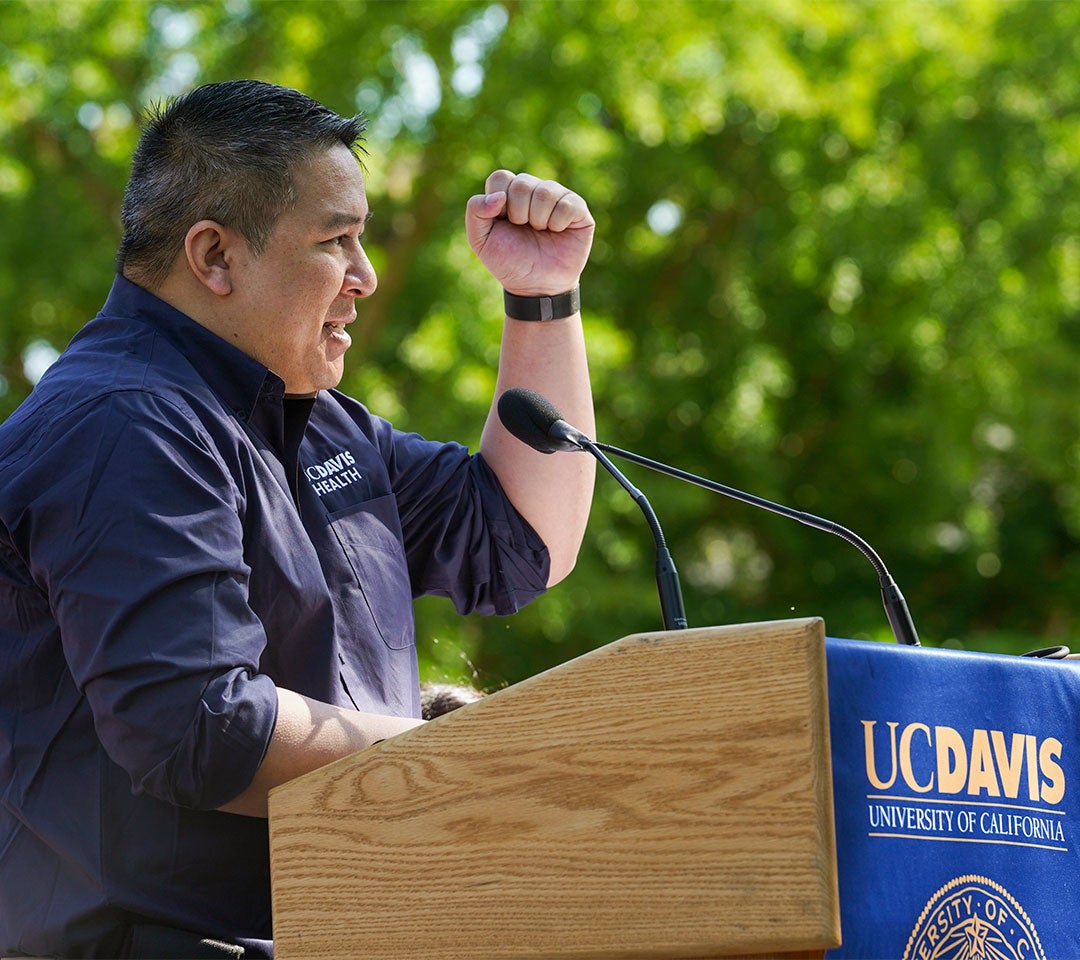 Dr. Nam Tran, in a UC Davis Health shirt, speaks at a podium with his fist raised at Picnic Day's opening ceremony, celebrating his role as both alum and 2022 Parade Marshal.