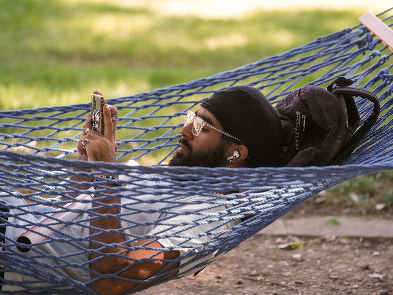 A UC Davis student scrolls on their phone while swinging in a blue hammock on campus.