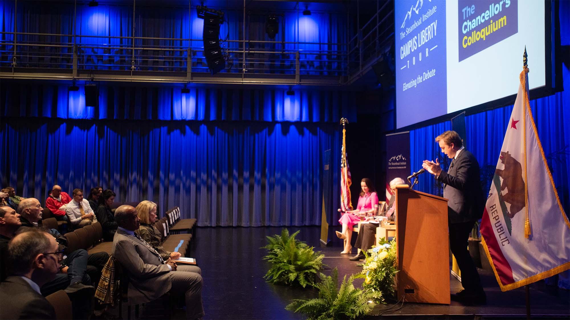 A speaker addresses an audience with blue lighting and a California flag in view.