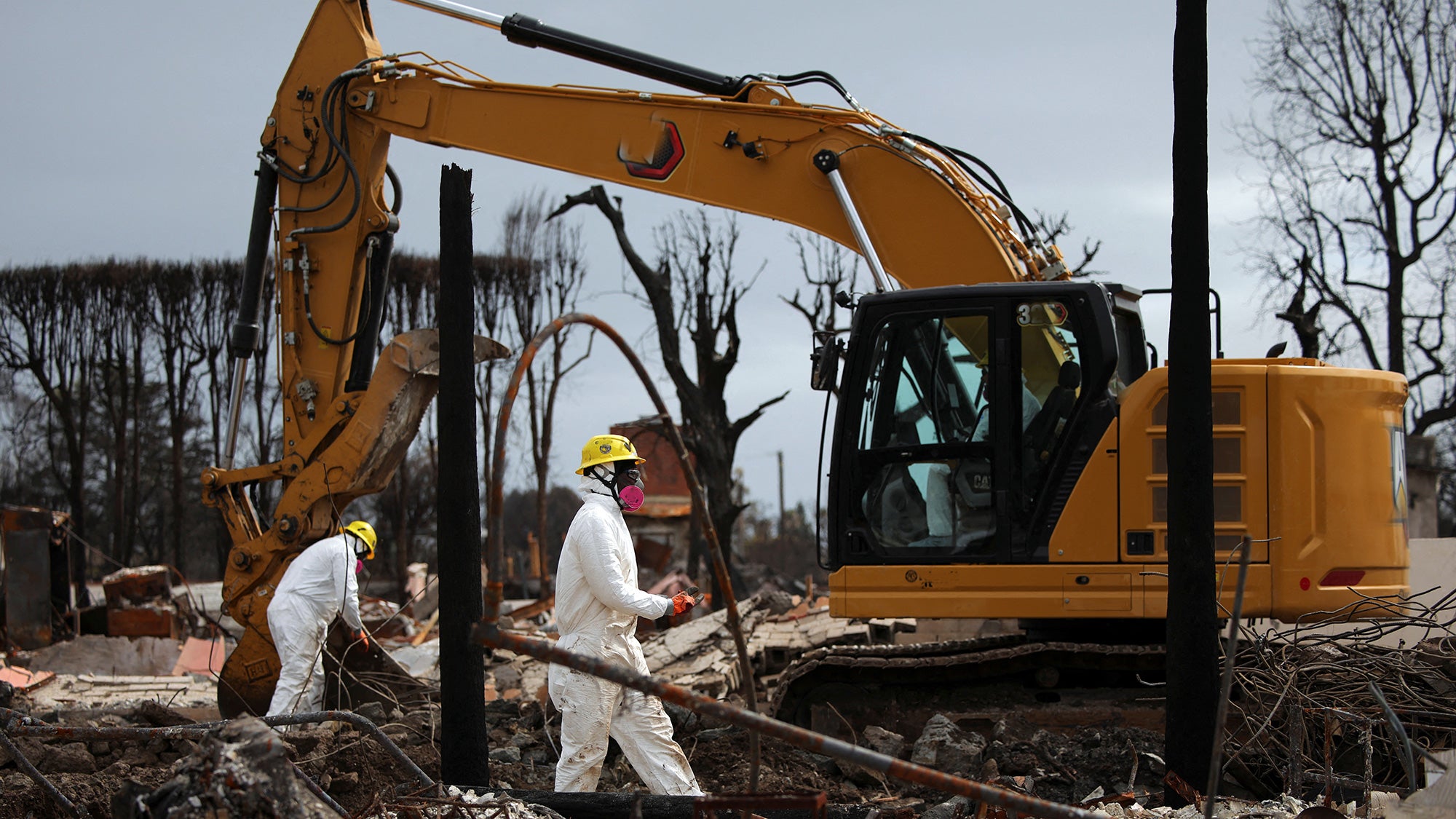 Two workers in white hazmat suits beside a yellow excavator at a muddy, bare-treed site