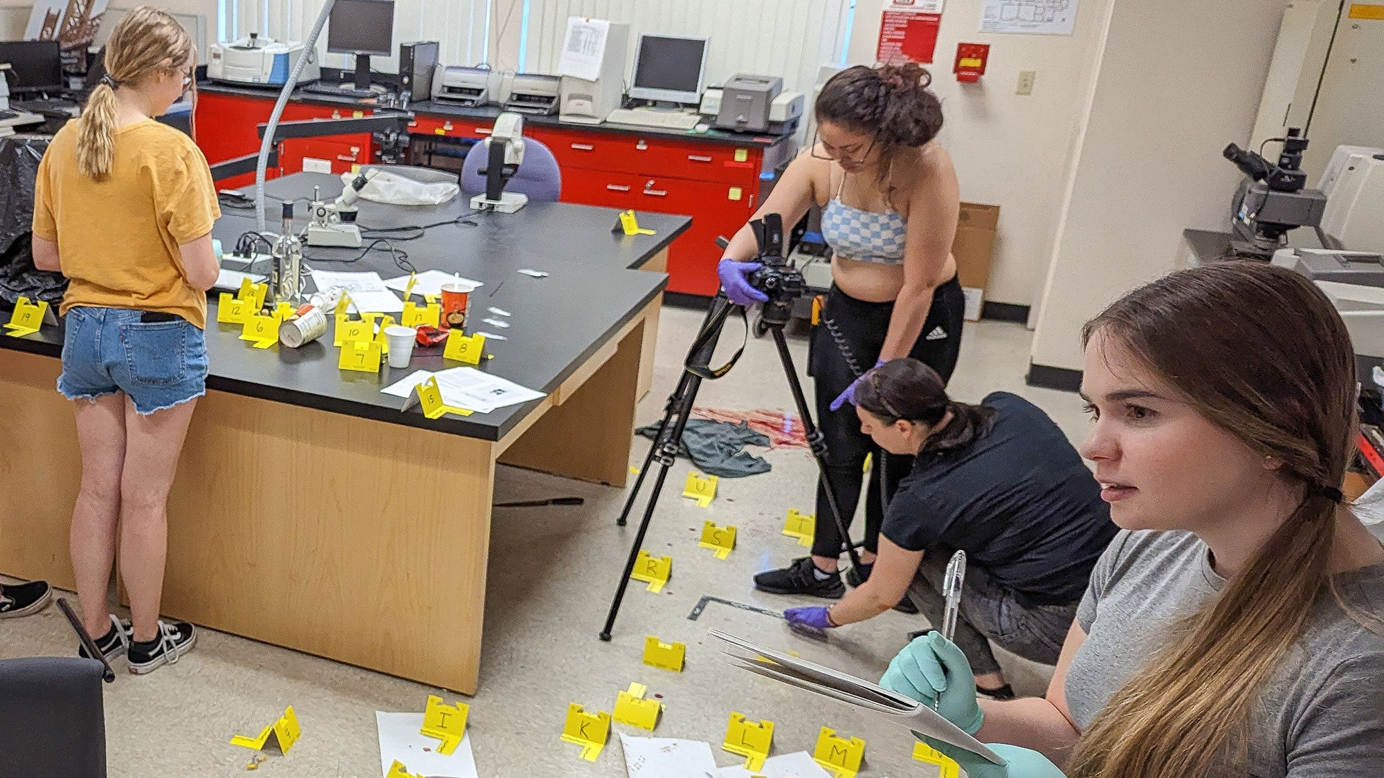 Students placing many yellow crime scene tags across a science lab bench while filming with a tripod.