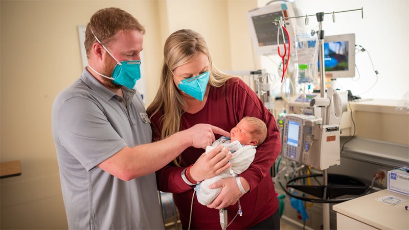 Masked parents in a hospital holding a swaddled newborn beside medical monitors
