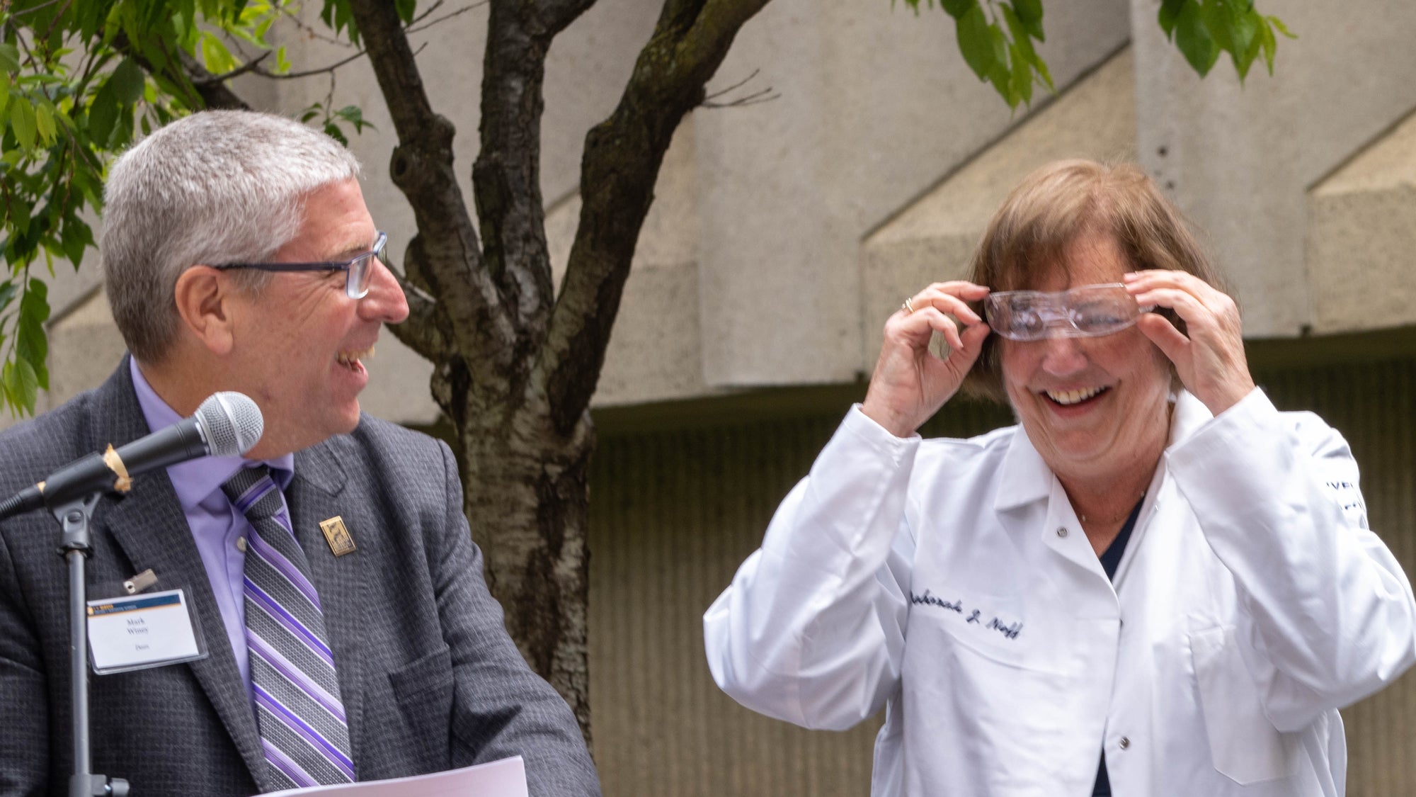 Woman in lab coat adjusts safety glasses, Dean Winey looks on from podium