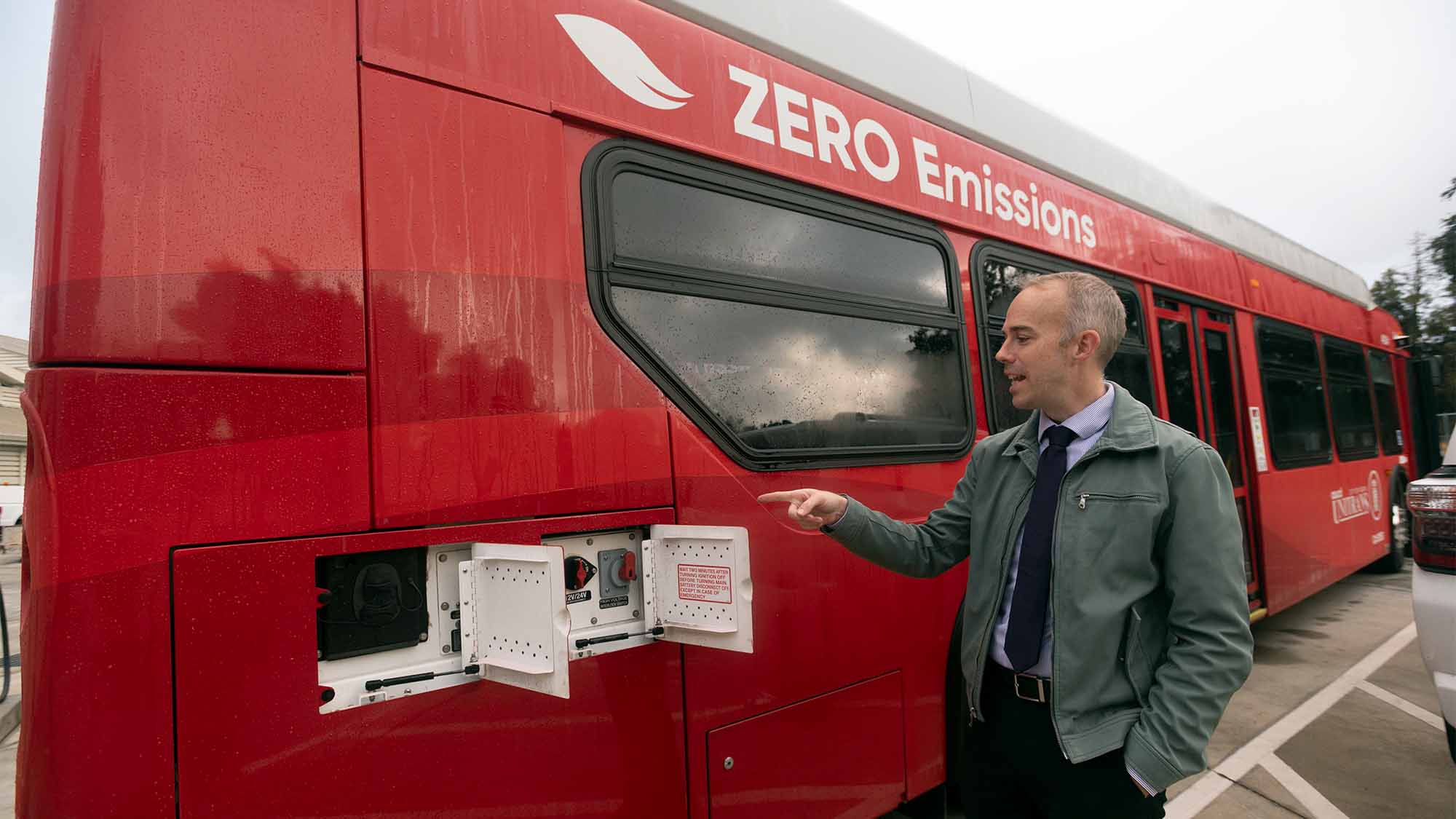 Man points to electric charging hook-ups on side of red Unitrans bus