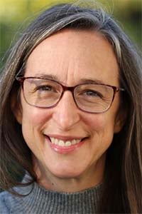 Close-up headshot of smiling woman with glasses, long brown hair, gray sweater, green background
