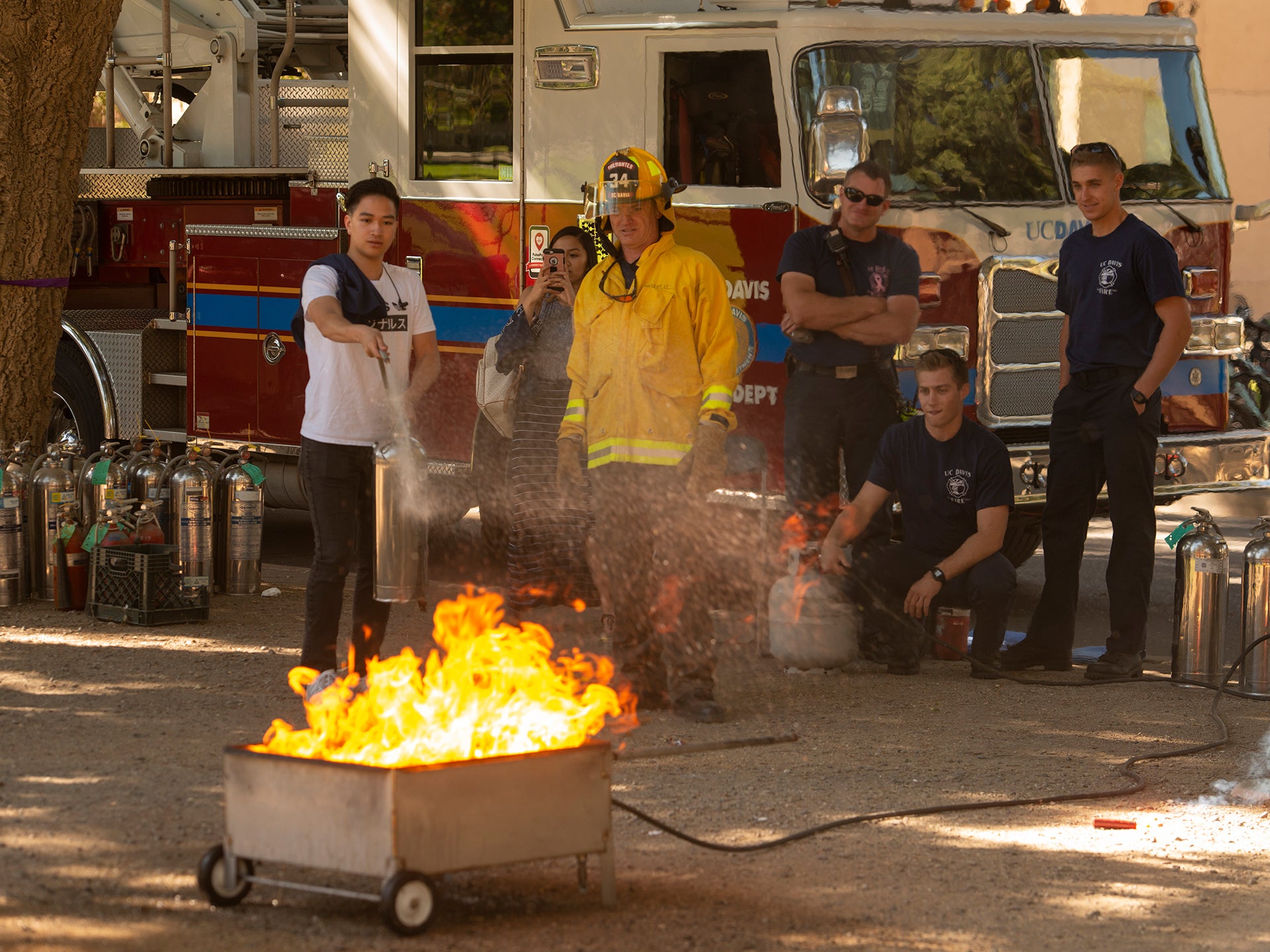 A UC Davis student extinguishes a bin fire while students and firefighters watch. A firetruck is in the background.