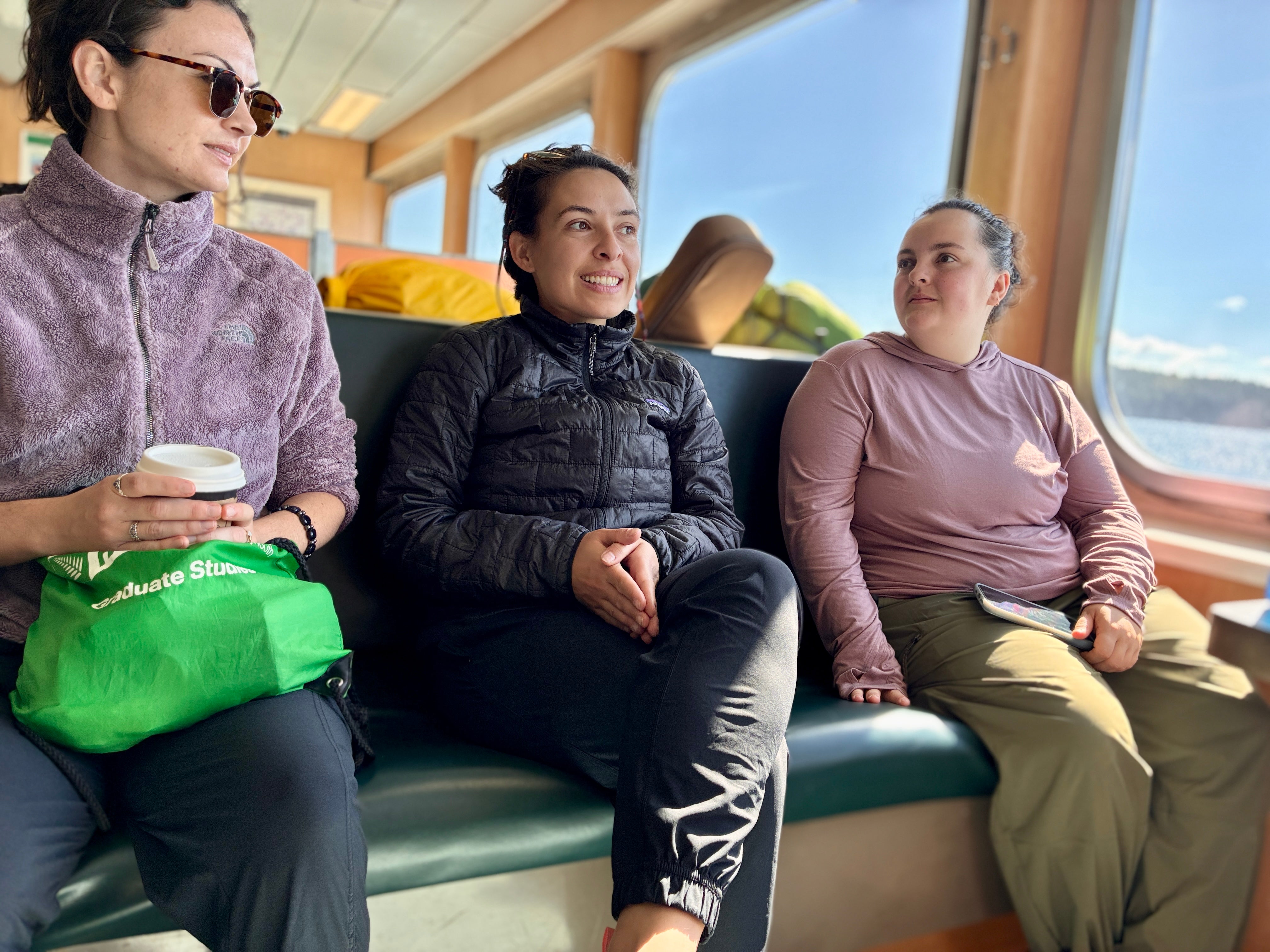 Three passengers sitting on a sunlit boat bench by large windows, chatting