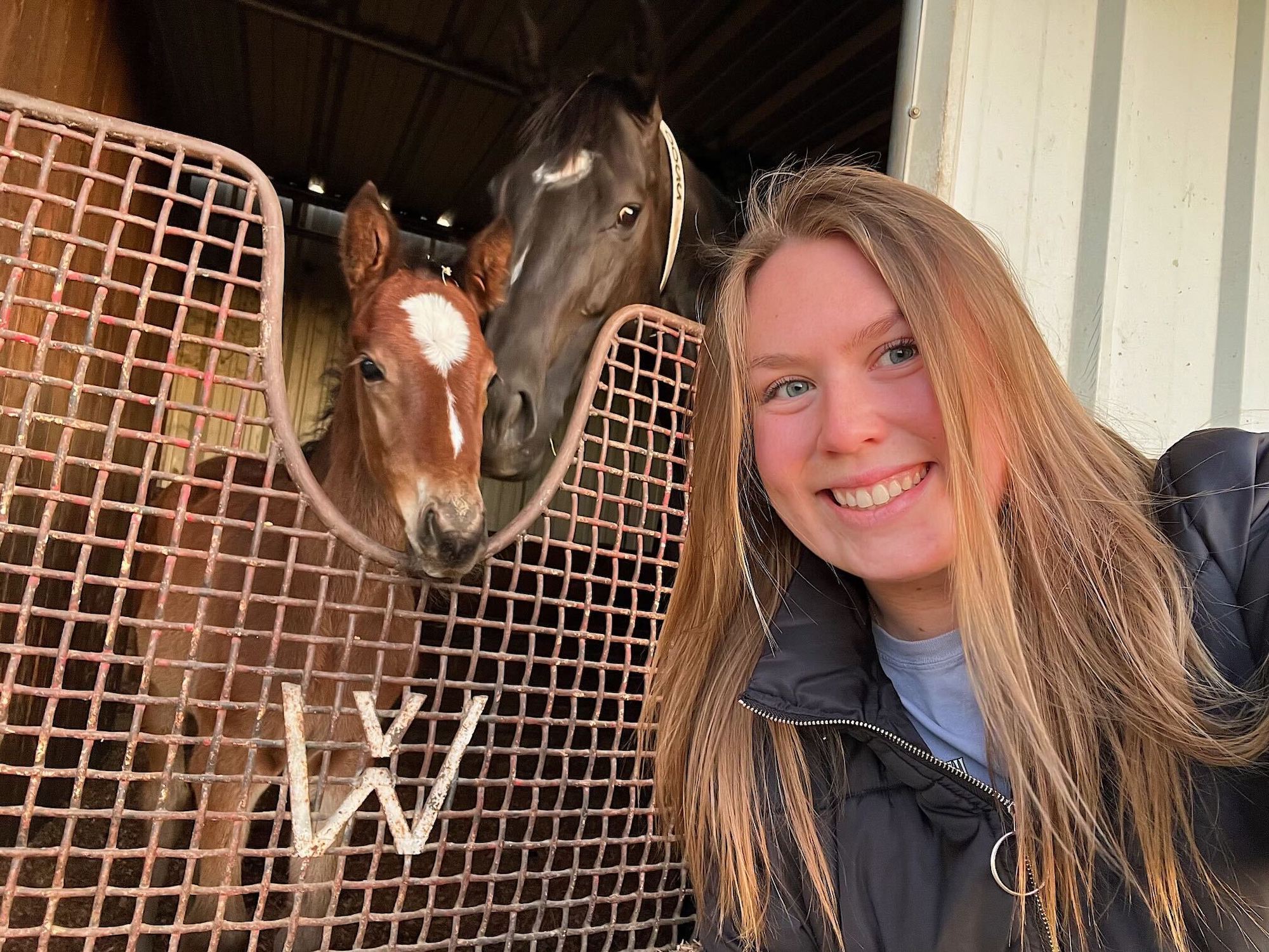A student poses for a selfie with two horses in a barn. 