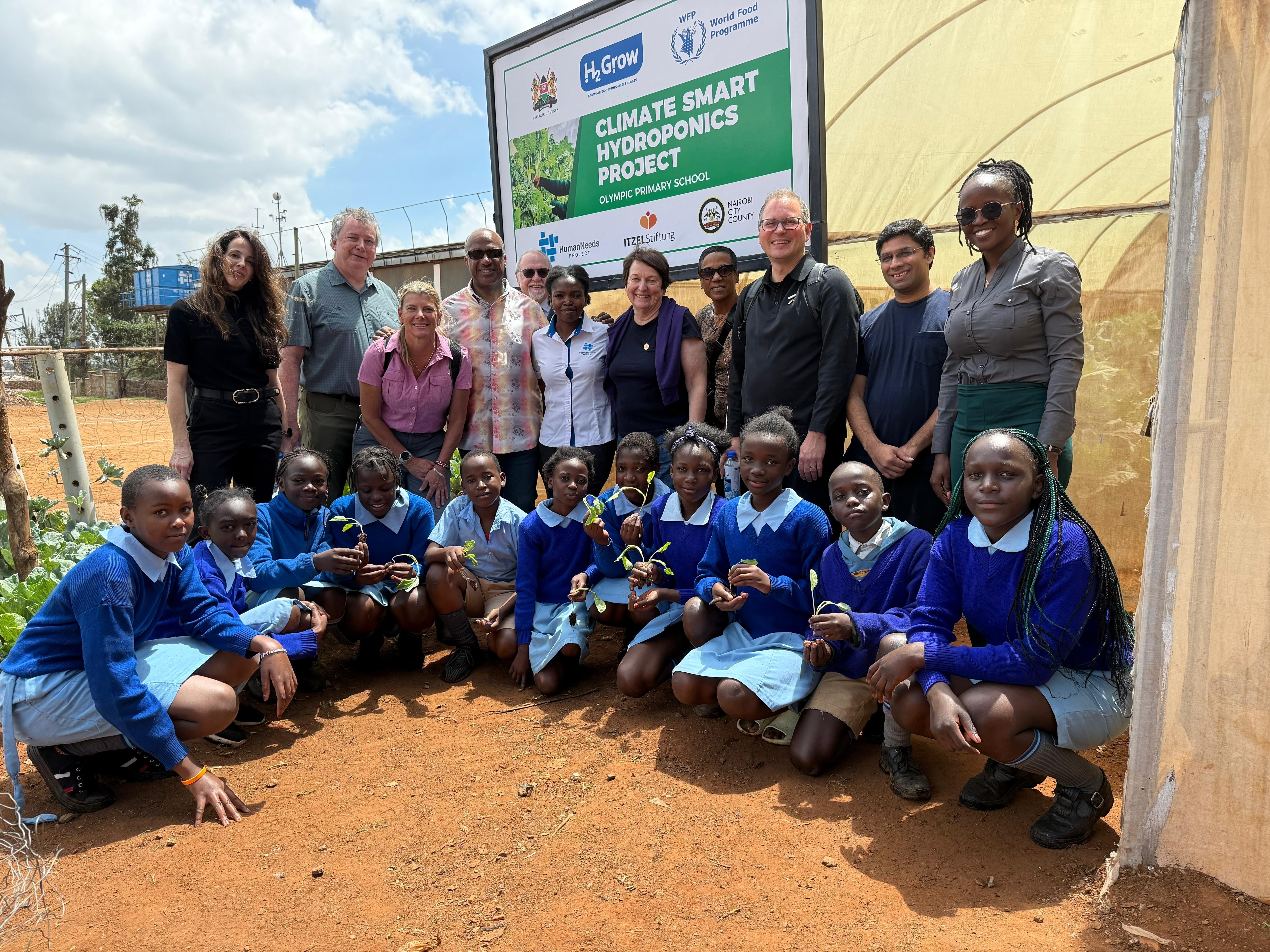 Group of 12 UC Davis representatives and partners stand behind 11 Kenyan children dressed in blue school uniforms,. A sign behind them reads Climate Hydroponics Project. 