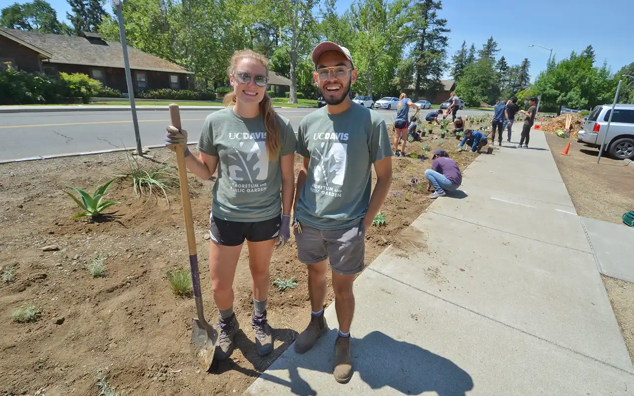 Two smiling students in green shirts on sidewalk during tree-planting