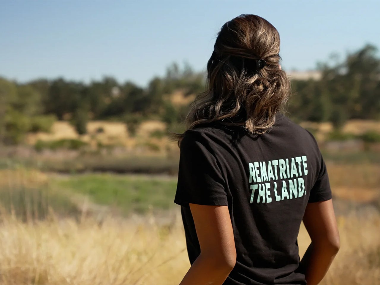 Melinda Adams looks out at a field. The back of her black shirt reads in light blue text: “Rematriate the land.”