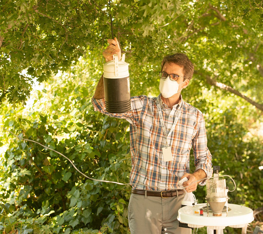 Professor Barker prepares a mosquito trap in a shaded outdoor area surrounded by greenery, contributing to UC Davis's research on mosquito-borne diseases.