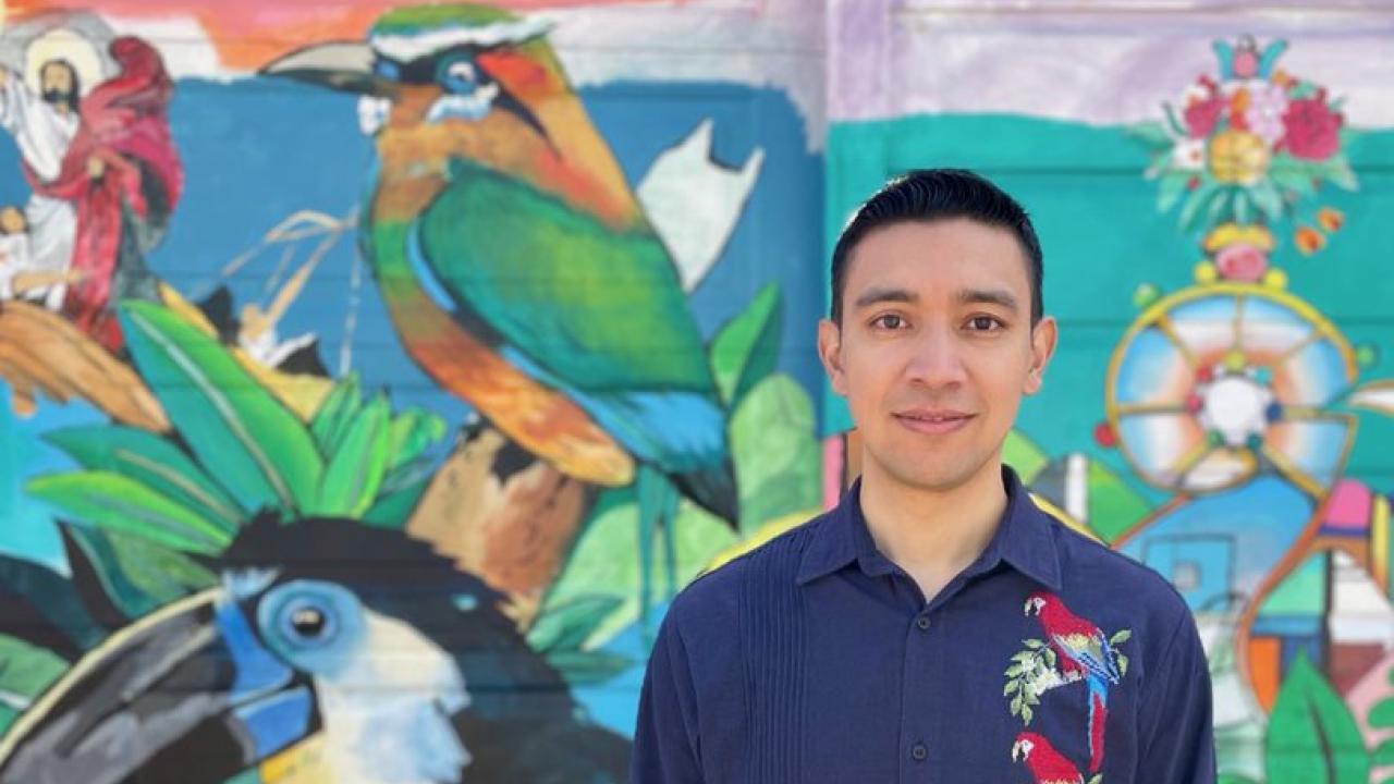 Mauricio Ernesto Ramírez in dark blue shirt with colorful embroidery on the left side, short dark hair looking directly at the camera in front of a colorful mural with people, green space and a mountain.
