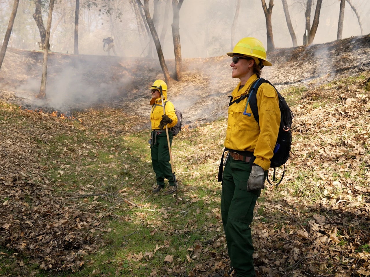 Yoshi Maezumi and Rebecca Wayman, in yellow firefighting jackets and helmets, stand in a meadow with light smoke in the background from a prescribed burn