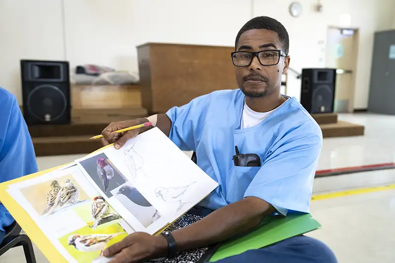 Man in blue scrubs and glasses seated indoors, holding open binder with photos and drawings