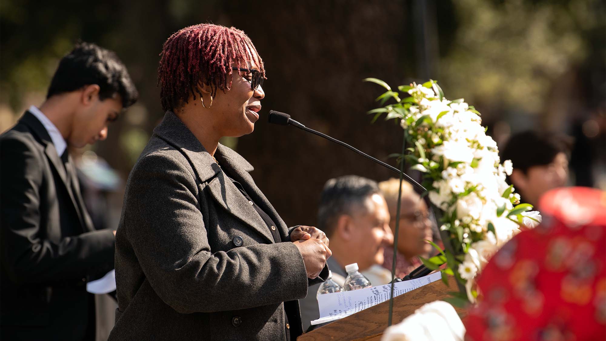 Quentisha Davis Wiles speaks at lectern, with wreath of white flowers in background.