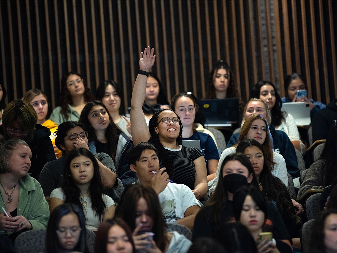 A student raises their hand and calls out a question from the back of the auditorium, surrounded by other students.