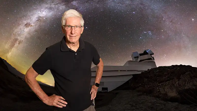 Older man with white hair and glasses in black shirt standing before observatory under Milky Way