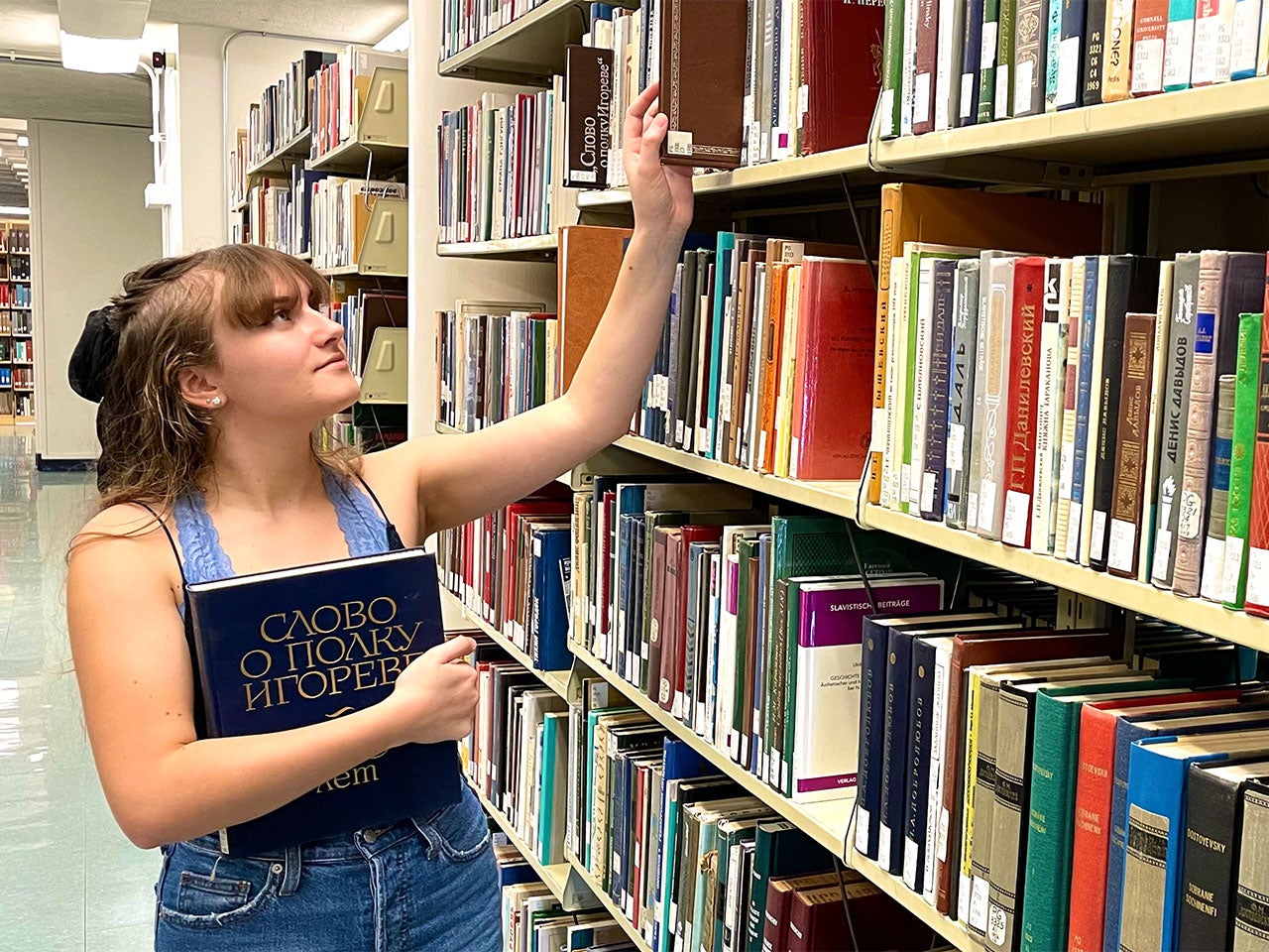 A shelf in Peter J. Shields Library displaying books with titles printed in Cyrillic. UC Davis Russian minor Ryleigh Praker reaches for a book while holding a blue book with a Russian title.