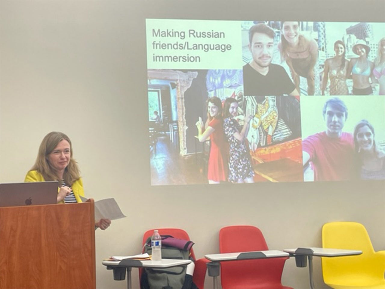 Professor Jenny Kaminer stands at a lectern in front of a projector screen. The screen displays a presentation with the title "Making Russian friends/Language immersion."