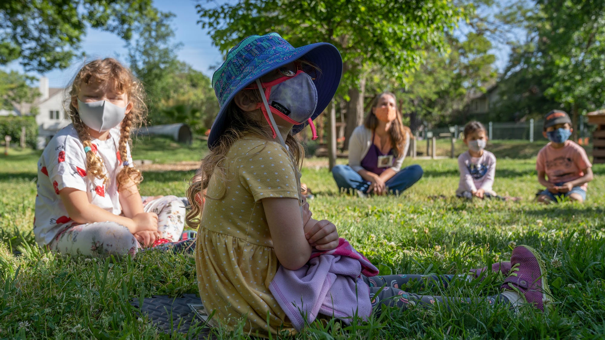 Children in masks, in circle, on grass, for outdoor lesson.